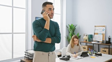 Telefondaki bir adam ve modern ofis ortamında parlak ışıklandırması ve temiz bir masa alanı olan odaklanmış bir kadın..