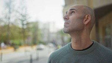 Yakışıklı, kel, sakallı ve küpeli adam işlek bir şehir sokağında düşünceli bir şekilde dikiliyor..
