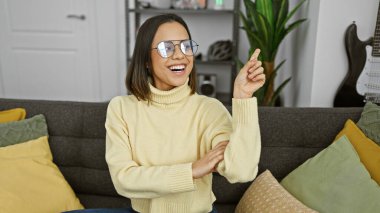 Gözlüklü, mutlu genç bir kadın. Oturma odasındaki koltukta oturuyor ve gülümsüyor..