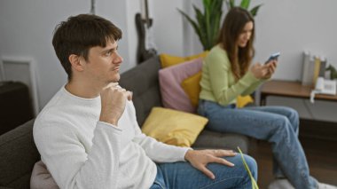 Akıllı telefon kullanan düşünceli bir adam ve kadın modern bir oturma odasındaki koltukta ayrı ayrı oturup ilişki sorunları tasvir ediyorlar..