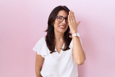 Orta yaşlı İspanyol kadın, günlük beyaz tişört ve gözlük takıyor. Bir gözünü eliyle kapatıyor. Yüzünde kendine güvenen bir gülümseme ve sürpriz bir duygu var.. 