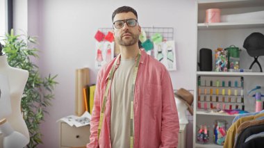 Kendinden emin genç bir adam canlı bir atölyede duruyor. Etrafı dikiş malzemeleri ve mankenlerle çevrili..