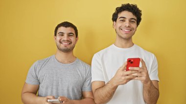 Sarı arka planda akıllı telefonları tutan iki gülümseyen İspanyol adam..