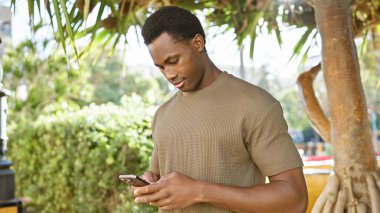 Genç, Afro-Amerikan bir adam güneşli bir parkta yemyeşil bir telefon kullanıyor.