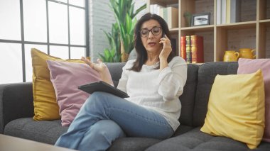 Olgun bir İspanyol kadın evinde elinde tabletle bir kanepede otururken akıllı telefonuyla konuşur..