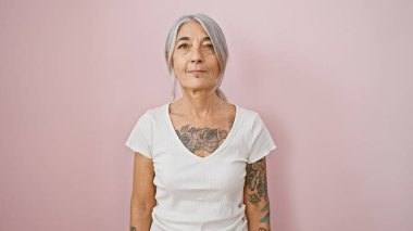 Ciddi, gri saçlı, orta yaşlı, spor kıyafetli bir kadın pembe izole bir arka plan üzerinde duruyor, spor salonunu kucaklıyor.