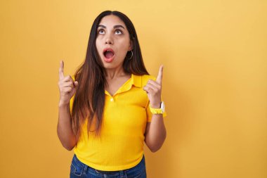 Sarı arka planda duran genç Arap kadın şaşırmış ve şaşırmış bir şekilde yukarı bakıyor ve parmaklarıyla işaret ediyor ve kollarını kaldırıyor.. 