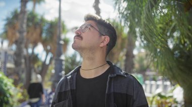 Bıyıklı İspanyol bir adam, güneşli bir parka dikkatlice bakar. Etrafı yeşilliklerle çevrilidir. Açık havada sakin bir ortam oluşturur..