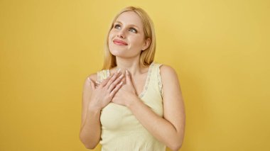 Grateful young woman with blonde hair and a heartfelt expression standing against a yellow backdrop clipart