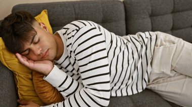 Attractive, young hispanic man found comfortable relaxation, sleeping soundly on the cozy sofa, tired and exhausted, lying in the comfortable ambience of his indoor home living room, simply unwinding. clipart
