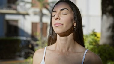 Genç ve güzel İspanyol kadın dengeyi ve özgürlüğü buluyor, temiz havayı soluyor, tamamen dalmış meditasyon yapıyor, şehirdeki güneşli yeşil parkta gözleri huzur içinde kapalı.