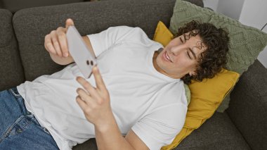 Kıvırcık saçlı genç bir İspanyol koltukta rahatlar, rahat bir oturma odasında telefonunu kullanarak gülümser..