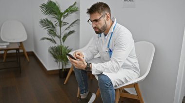 Yakışıklı bir İspanyol genç adam, odaklanmış bir doktor, klinik bekleme odasında otururken akıllı telefonuyla mesaj atıyor, işine dalmış, modern tıp profesyonelliğinin bir portresi..