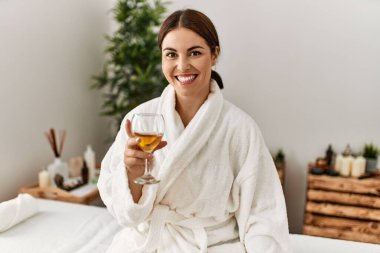 Young beautiful hispanic woman wearing bathrobe drinking wine at beauty salon