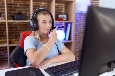 Video oyunları oynayan beyaz bir kadın, kulaklık takıyor ve el parmaklarıyla reklamını yapıyor, ciddi ve sakin bir yüz ifadesi var. 