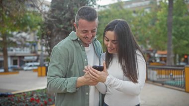 Neşeli bir çift bir anı paylaşıyor. Şehir parkında birlikte akıllı telefona bakıyorlar. Etrafı yeşilliklerle çevrili..