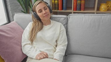 Rahat genç bir kadın, rahat bir oturma odasındaki kanepede otururken kulaklıkla müzik dinlemekten hoşlanır..