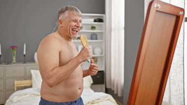 Keyifli, üstsüz, son sınıf öğrencisi rahat bir yatak odasında aynanın önünde saç fırçasıyla şarkı söylüyor..