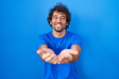 Hispanic young man standing over blue background smiling with hands palms together receiving or giving gesture. hold and protection 