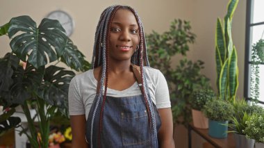 Kendine güvenen, saçları örgülü bir Afrikalı Amerikalı kadın bir çiçekçide duruyor. Etrafı yeşilliklerle çevrili, güzel ve özgüvenli..