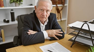 Ciddi bir kıdemli adamın büyüleyici portresi, başarılı bir iş adamı, ofiste odaklanmış bir bakışla oturuyor, kollarını kavuşturuyor. İş yerinde olgun bir özgüvenin örneği.. 