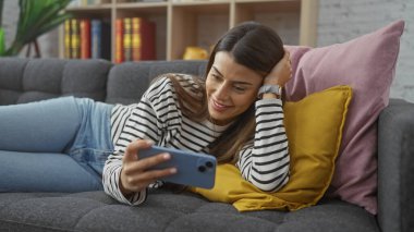 Gülümseyen İspanyol kadın, elinde akıllı bir telefonla kanepede uzanıyor, rahat ve rahat bir atmosfer yayıyor.