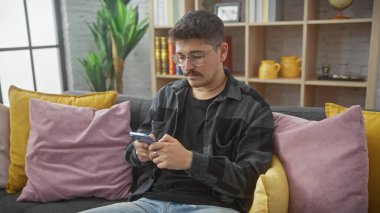 Bıyıklı, akıllı telefon kullanan, sarı kanepede oturan, mor yastıklı genç bir İspanyol..