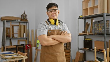 Confident young asian man with crossed arms wearing safety gear in a well-organized carpentry workshop clipart