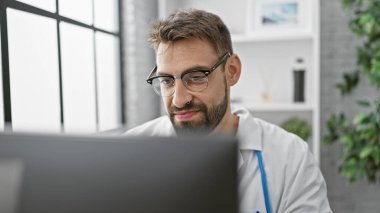 Sakallı, genç, İspanyol bir doktor, bir klinikte bilgisayarını kullanıyor.
