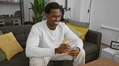 Gülümseyen genç siyah adam modern bir oturma odasında İsveç kronu notları sayıyor.