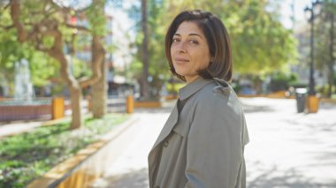 Olgun bir İspanyol kadın, güneşli bir parkta kendine güvenerek durur, zarafet ve duruş yayar..