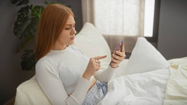 İyi ışıklandırılmış bir yatak odasında bir yatakta otururken akıllı telefon kullanan genç bir kadın..