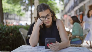 Gülümseyen güzel İspanyol kadın, kafedeki terasta otururken neşeli bir şekilde akıllı telefon kullanıyor, kahve içiyor, dijital dünyayla iletişim kuruyor, mutluluğunu arttırıyor..