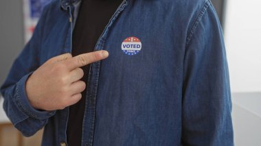 Caucasian man in office pointing at 'i voted' sticker on denim shirt near american flag clipart