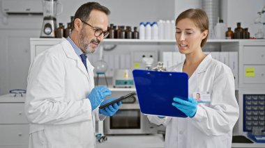 Laboratuvardaki iki memnun bilimadamı, bir dokunmatik dokunmatik yastıkta önemli bir belgeyi okurken, yoğun bir tıbbi araştırma oturumundan sonra sıcak bir anlaşma tokalaşması yaşadılar..