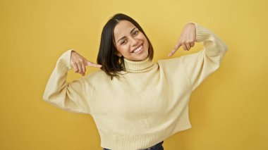 Krem rengi süveter giyen gülümseyen genç İspanyol kadın sarı arka planda poz veriyor, kendine güven ve neşe saçıyor..