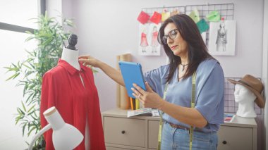 Tabletli olgun bir İspanyol kadın terzi, tasarım çizimleri ve kırmızı giysili bir mankenle çevrili iyi örgütlenmiş bir atölyede zanaata girişiyor..