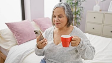 Gülümseyen olgun bir kadın, rahat yatak odasında rahatça otururken akıllı telefonuyla sabah kahvesinden zevk alır..