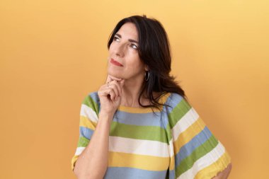 Middle age hispanic woman standing over yellow background with hand on chin thinking about question, pensive expression. smiling with thoughtful face. doubt concept. 