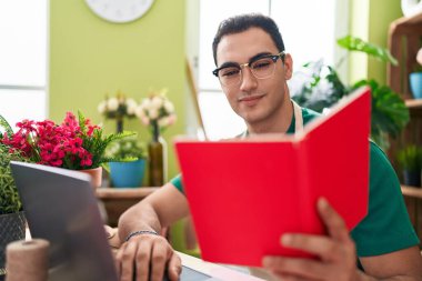 Young hispanic man florist using laptop reading book at flower shop