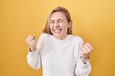 Sarı arka planda beyaz süveter giyen beyaz bir kadın çok mutlu ve heyecanlı bir şekilde kollarını kaldırıp, gülümseyerek ve başarı için çığlık atarak kazanan bir jest yapıyor. kutlama konsepti. 