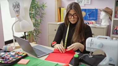 İyi donanımlı bir atölyede kumaşla çalışan, etrafı dikiş aksesuarlarıyla çevrili odaklanmış genç bir kadın terzi.