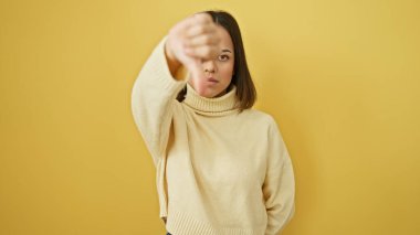 Nötr yüzlü İspanyol genç kadın sarı duvara karşı kazak giyip baş parmak işareti yapıyor..