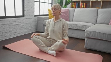 Rahat olgun bir kadın, gri bir kanepe ve bir kitaplığın yer aldığı huzurlu bir evde pembe bir yoga minderi üzerinde meditasyon yapar..