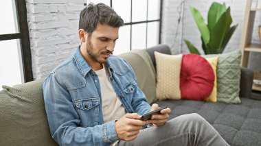 Yakışıklı, genç İspanyol bir adam rahat bir şekilde evdeki bir kanepede rahatlar, akıllı telefonunu güzel dekore edilmiş bir oturma odasında gezer..