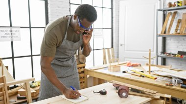 Genç bir adam atölyede çalışıyor, güvenlik gözlüğü ve önlük takıyor, not alıyor ve telefonla konuşuyor..