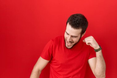 Gündelik kırmızı tişört giyen genç İspanyol adam mutlu ve neşeli dans ediyor, gülümsüyor rahat ve kendinden emin bir şekilde müzik dinliyor. 