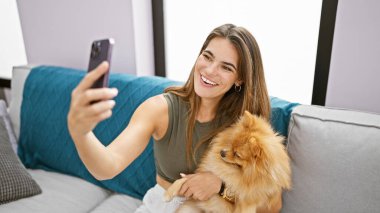 Neşeli, genç İspanyol kadın evinde, oturma odasındaki kanepesinde rahatça gülümsüyor. Evcil köpeğiyle mutlu bir selfie çekmek için akıllı telefon teknolojisi kullanıyor..