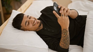 Kendine güveni tam genç Latin bir adam, evinde rahat bir yaşam tarzının portresi olan, yatağında rahat bir şekilde yatarken akıllı telefonuyla gülümseyip mesaj atıyor.