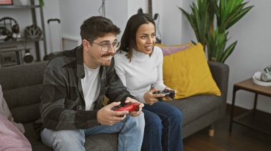 Gülümseyen bir erkek ve kadın, rahat, modern bir oturma odasındaki kanepede video oyunları oynuyorlar..